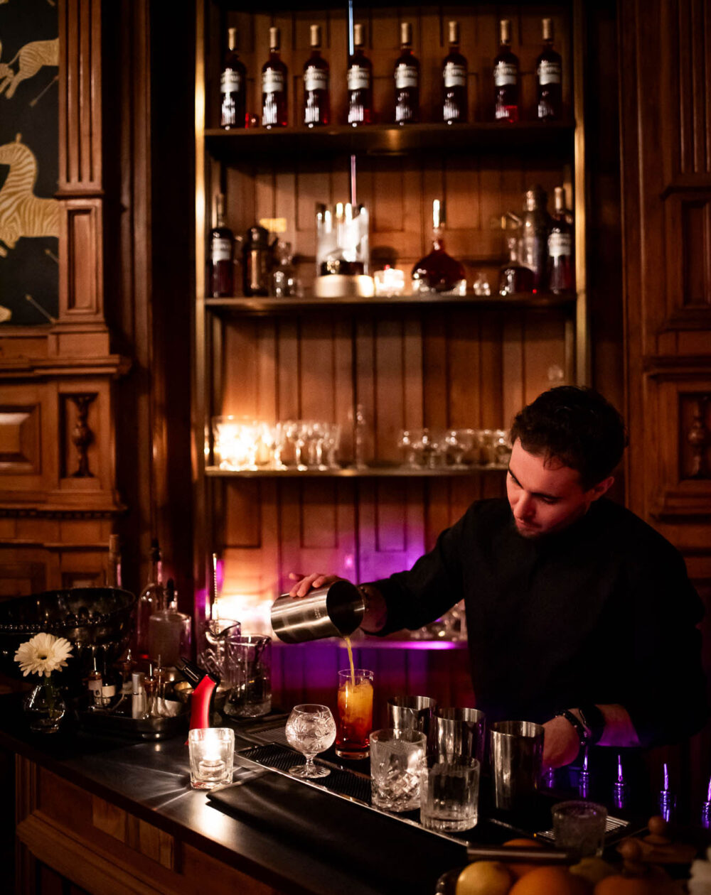 Bartender pouring a cocktail from a shaker at a dimly lit bar with illuminated shelves of spirits and candles