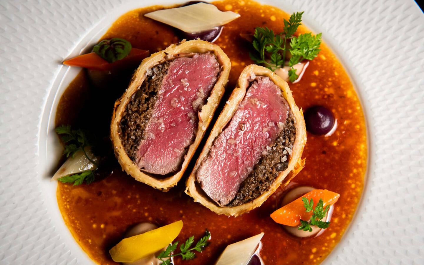 Two slices of beef Wellington showing medium-rare beef and mushroom duxelles in pastry, plated with root vegetables and rich sauce
