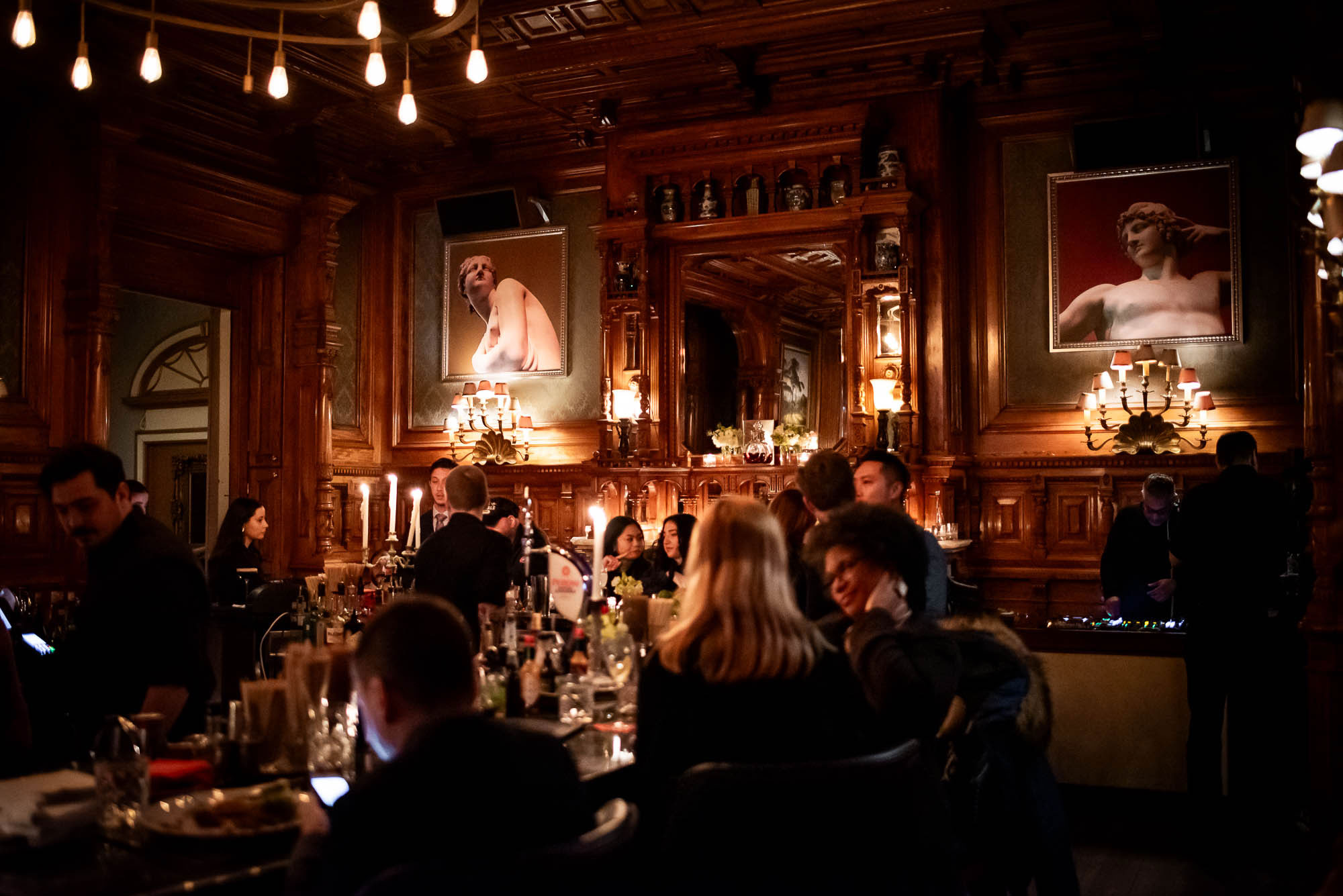Elegant restaurant dining room with dark wood paneling, ornate mirrors, classical portrait paintings, and Edison bulb lighting, filled with dinner guests