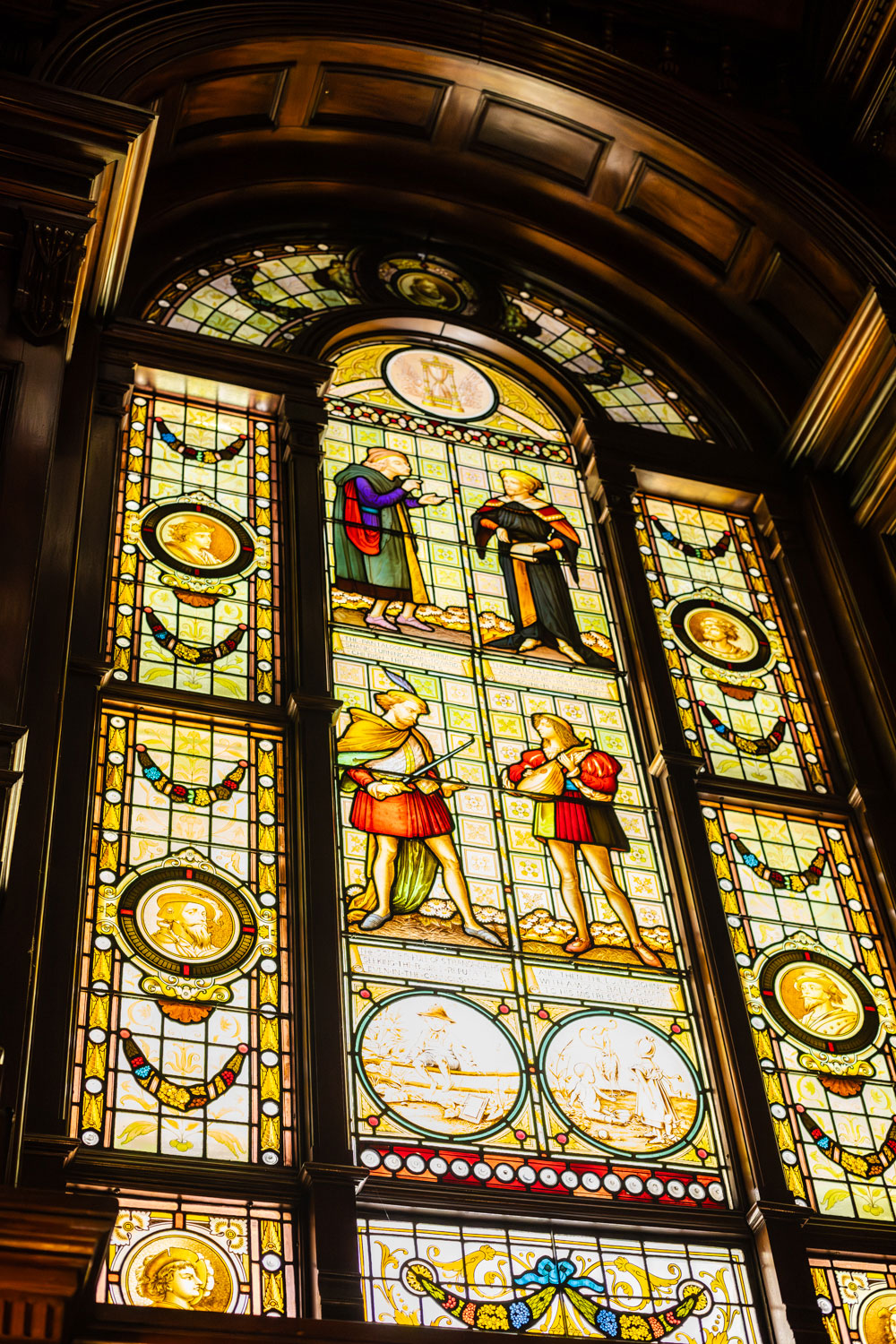 Ornate Victorian stained glass windows with historical figures and portrait medallions, set in dark wood archway