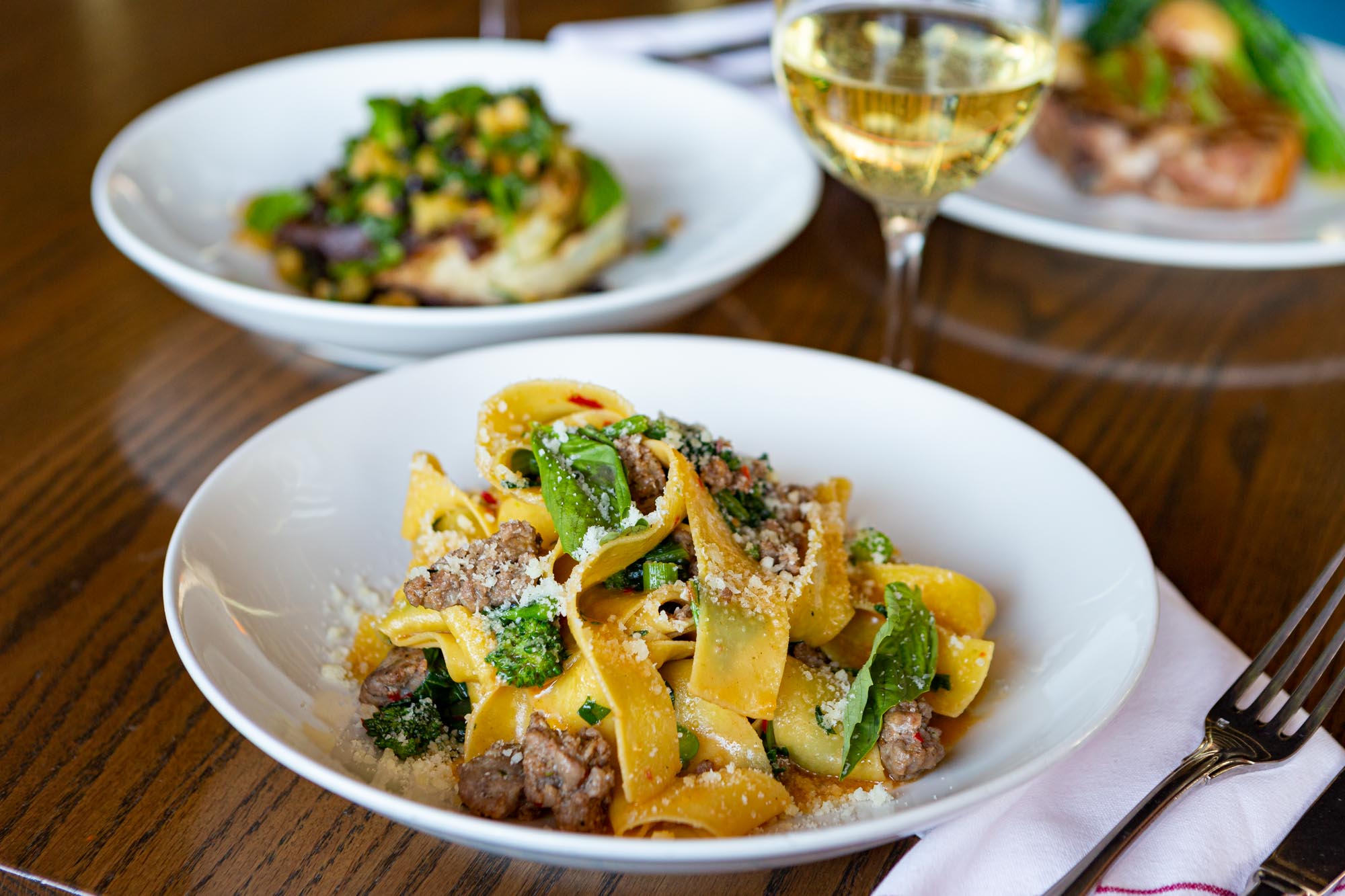 Pork Pappardelle Pasta in a white bowl with other pasta dishes and a glass of wine