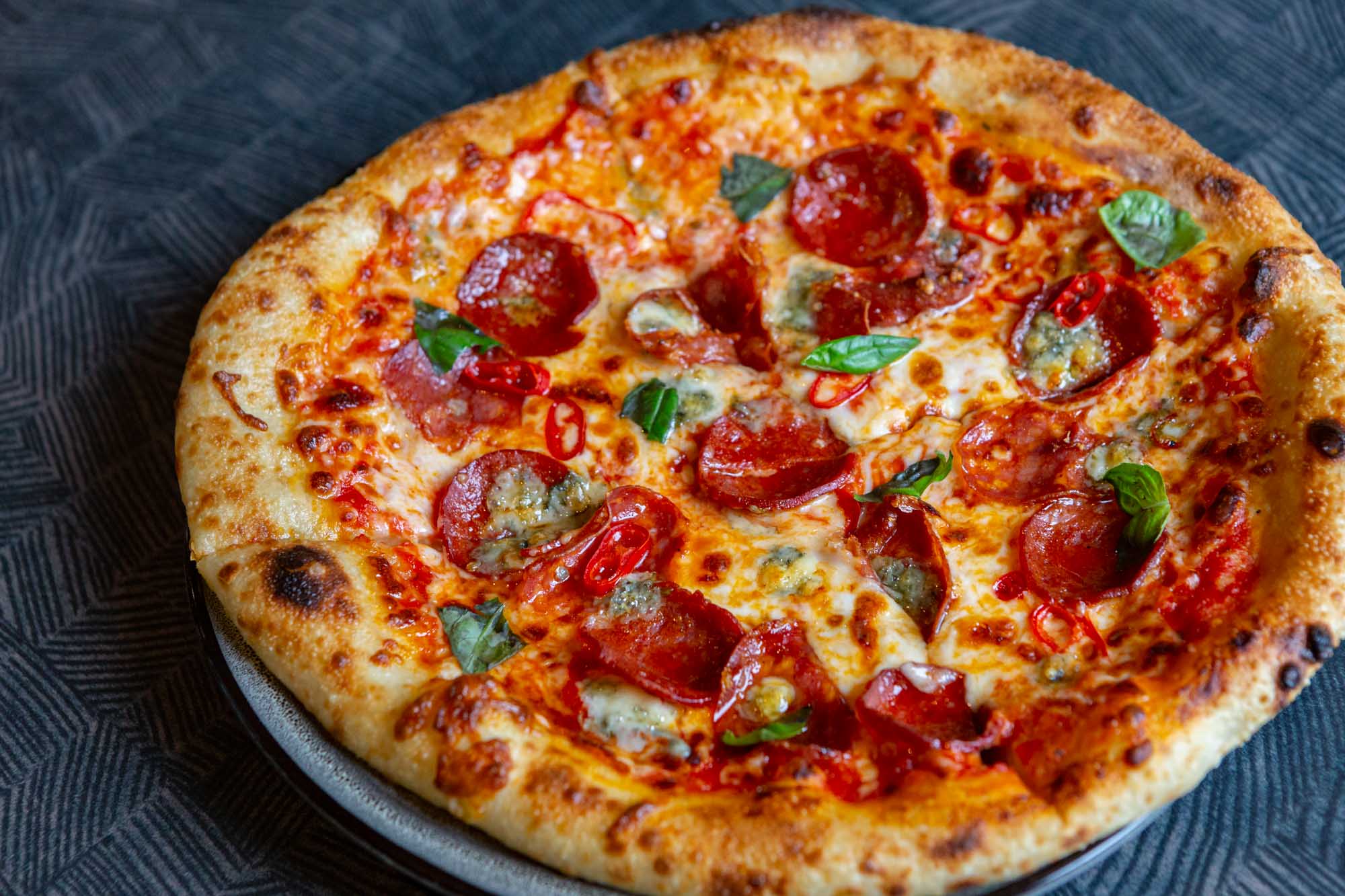 Top-down, slightly angled shot of a fire-roasted pizza on a plate with a dark grey background. The pizza has pepperoni and basil leaves and looks a bit crispy.