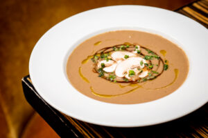 Top-down shot of Jump's Wild & Tame Mushroom Soup, a nice tan colour with artful plating and garnishes. The bowl is white and it's on a brown wooden table.