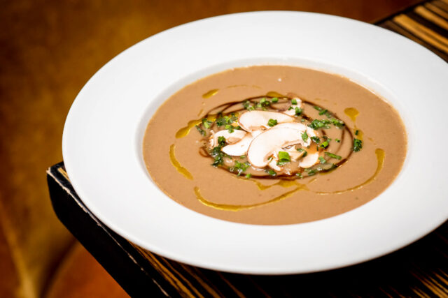 Top-down shot of Jump's Wild & Tame Mushroom Soup, a nice tan colour with artful plating and garnishes. The bowl is white and it's on a brown wooden table.