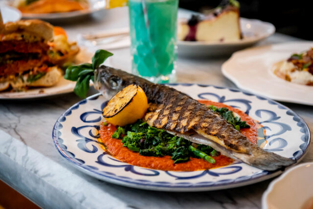 grilled whole branzino with a half roasted lemon on an ornate plate with other dishes in the backdrop