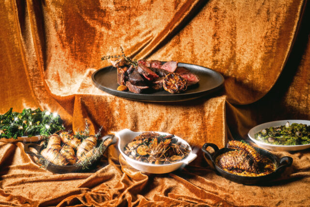 many dishes including steak on an elevated platform in front of a golden fabric