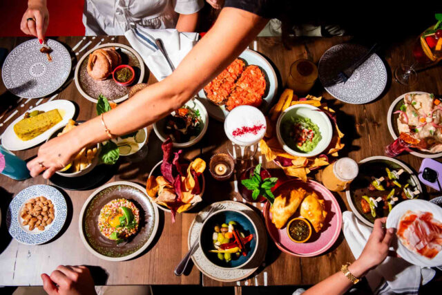 many hands reach for various latin-inspired dishes on a dark wooden table
