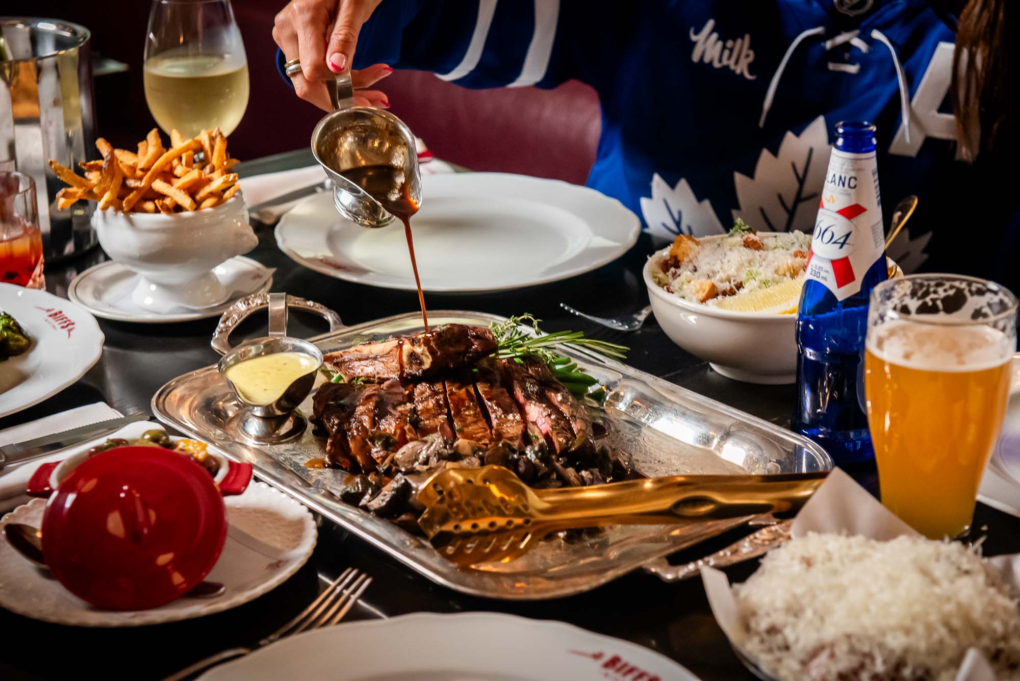 A group dining with a spread of dishes including a large cote de boeuf for two and a gravy boat being poured over top.