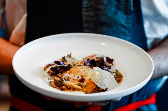 Person in blue apron with red straps hold plate with duck egg yolk ravioli with wiseats oyster mushrooms, ricotta, brown butter sauce and sylvan star grizzly gouda at Buffo