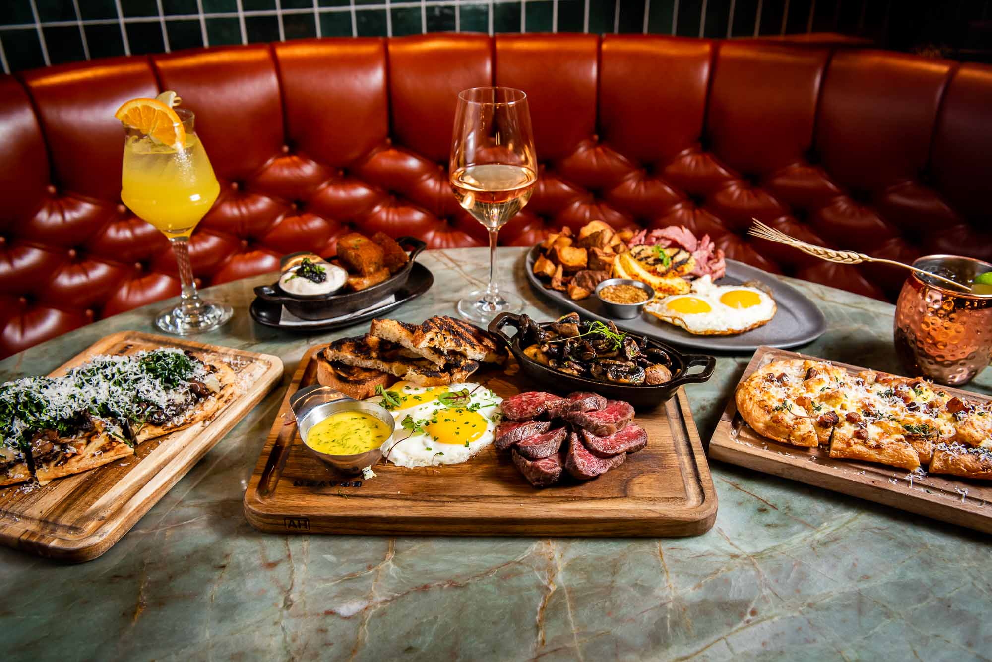 A full spread of Braven's breakfast items on a table with two full wine glasses. The background is a curved orange-brown leather sofa.