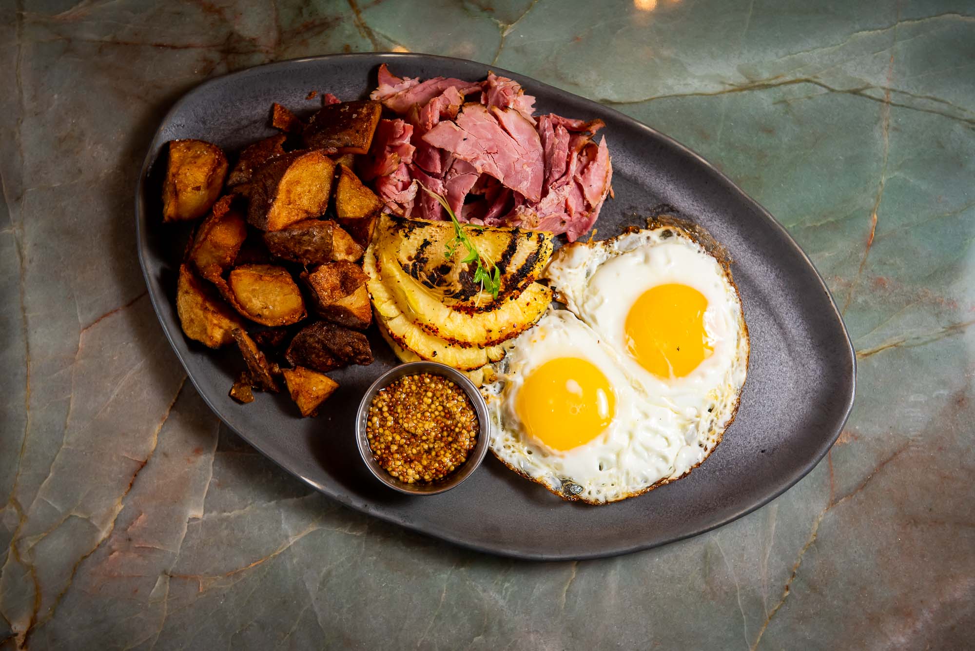 Top-down shot of fire-roasted ham and eggs on an oval-shaped grey plate and marble-like table.