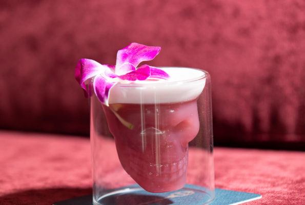 Dark pink cocktail in a skull-shaped glass, garnish with an orchid on a pink velvet background