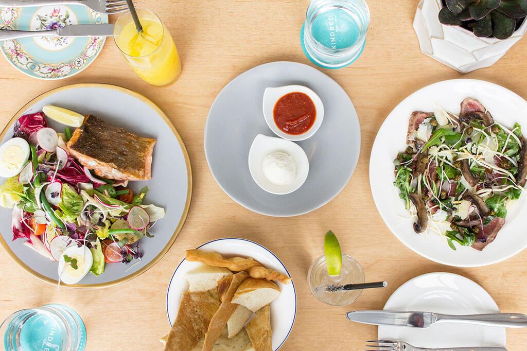 A spread on a wooden table at Kindred including the Salmon Nicoise Salad and the Alberta Beef Carpaccio, with condiments and other cutlery surrounding.