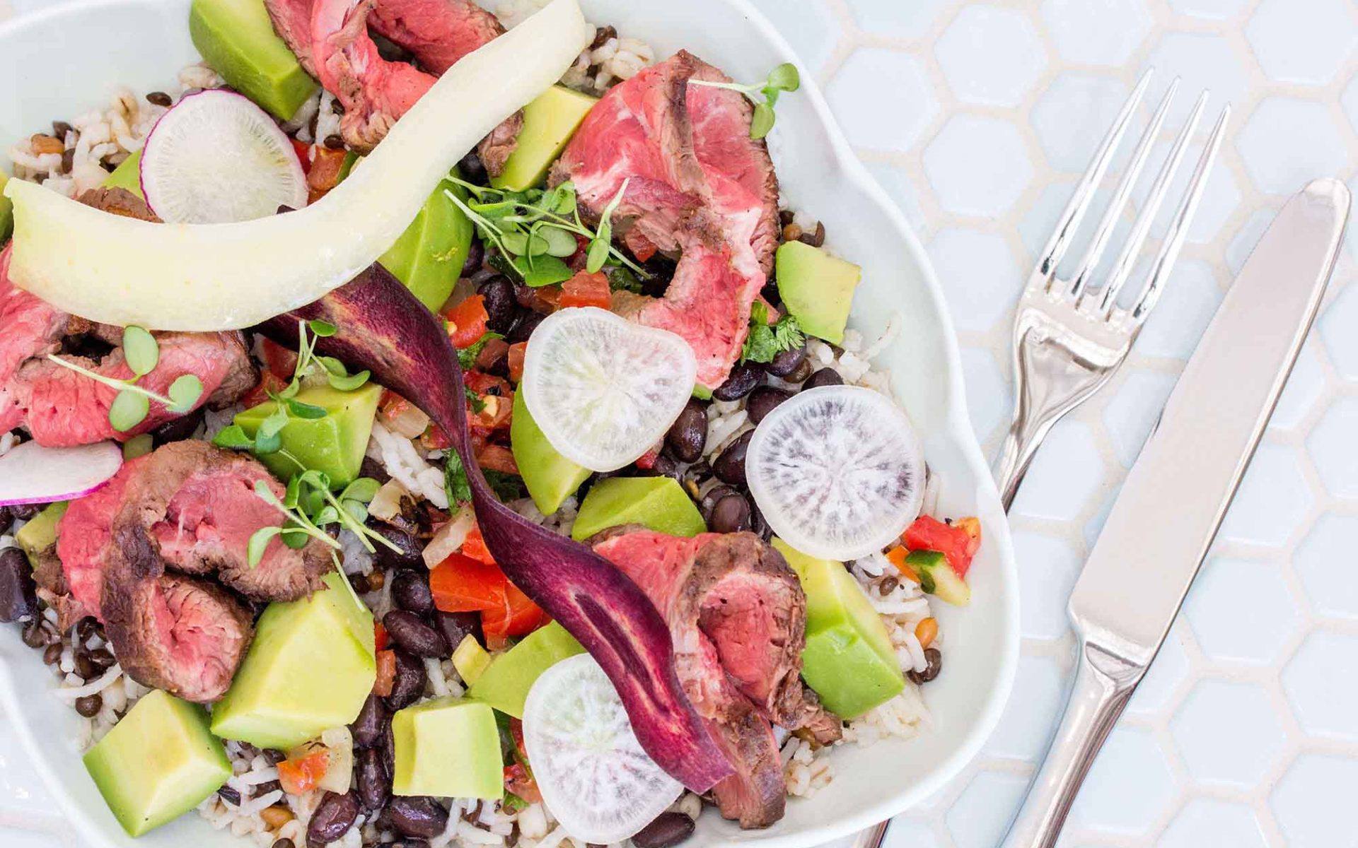 overhead of steak and grain bowl with slices of radish and avocado accompanied with knife and fork