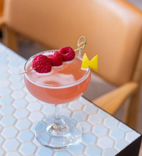 peach-coloured cocktail with raspberry garnish and lemon peel