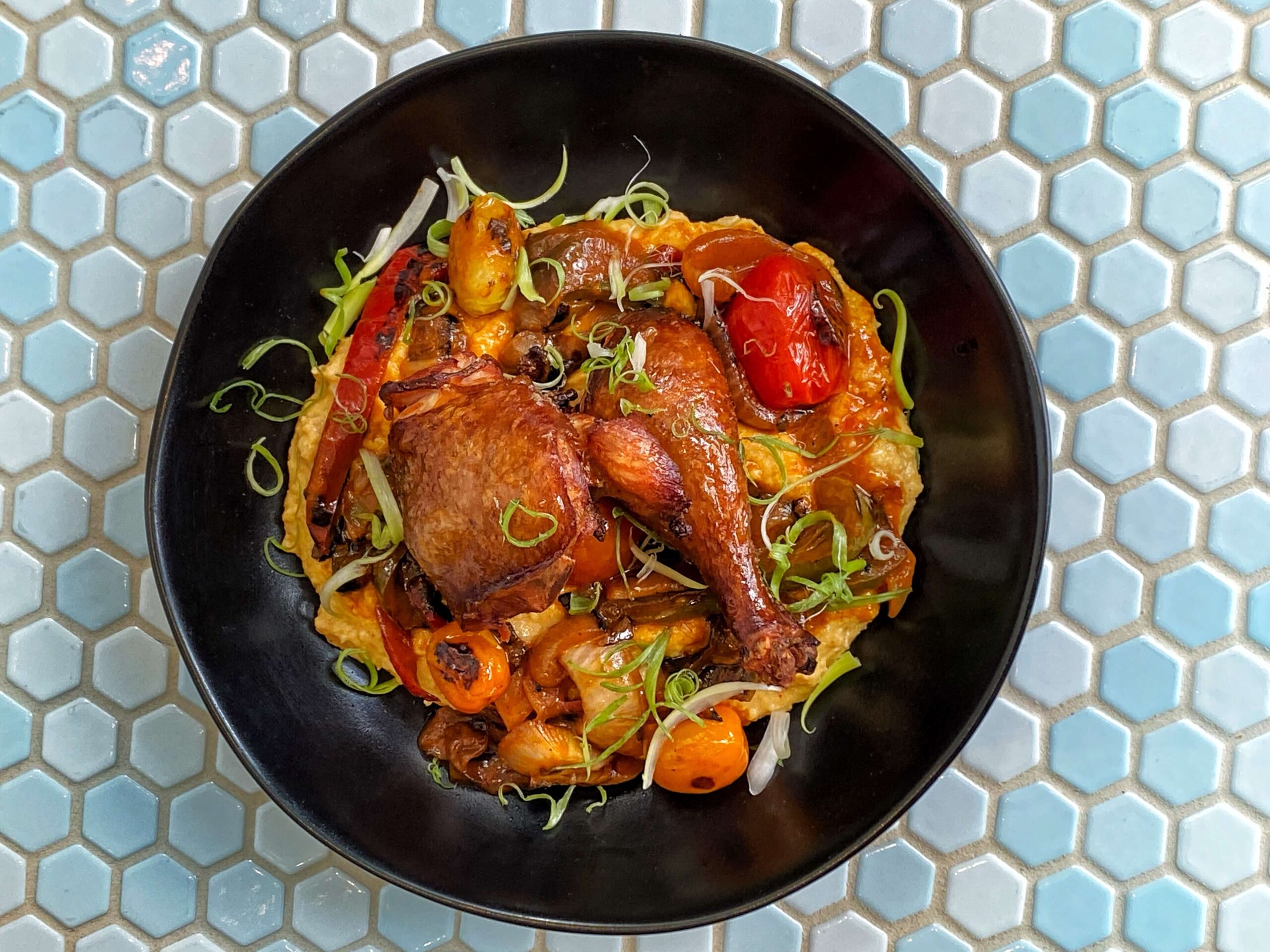 Roasted Chicken and Grits smoked legs with disco grits, nasty Buffalo piperade in black bowl on tiled blue table at Kindred Restaurant Edmonton