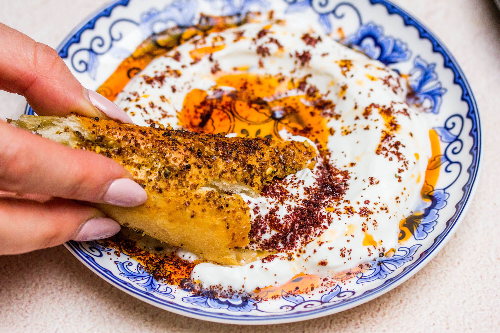 Hand with nail polish dipping bread into a blue and white bowl of labneh