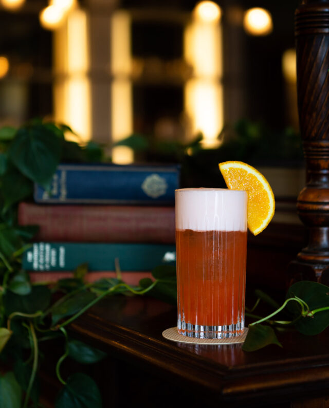 A bright orange cocktail garnished with an orange sitting in front of a stock of books