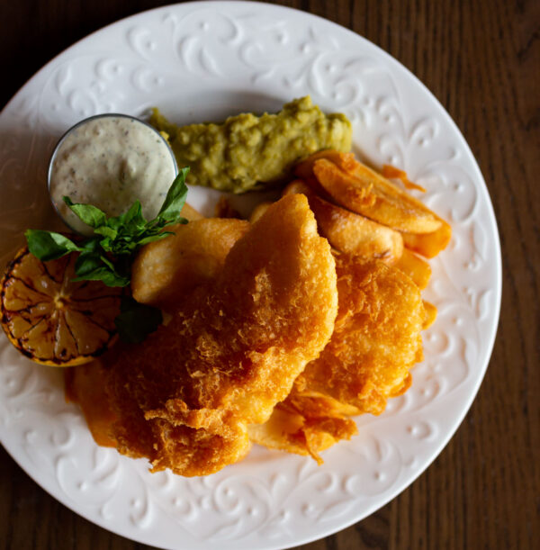 Ale Battered Halibut & Chips on a white plate with a side of tartar sauce, mushy peas and a grilled lemon half
